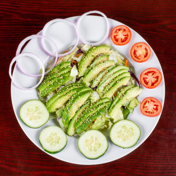 Avocado salad with lettuce, sesame seeds, red onion, tomato, and cucumber slices on a white plate.
