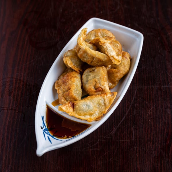 Crispy fried dumplings in a white boat-shaped dish with soy sauce on a dark wooden table.