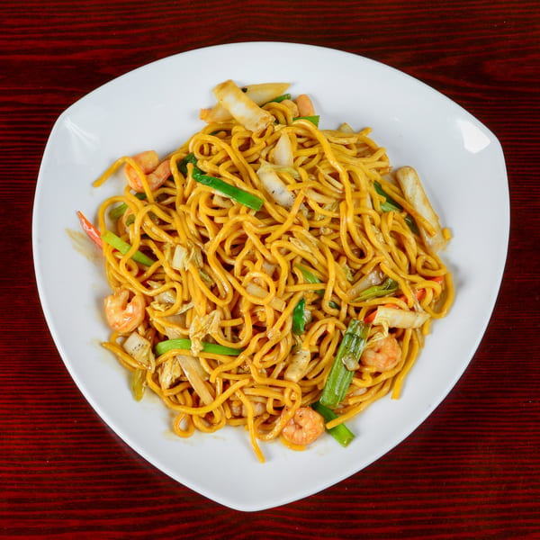 A white plate filled with golden noodles, shrimp, and mixed vegetables on a dark red wooden table.
