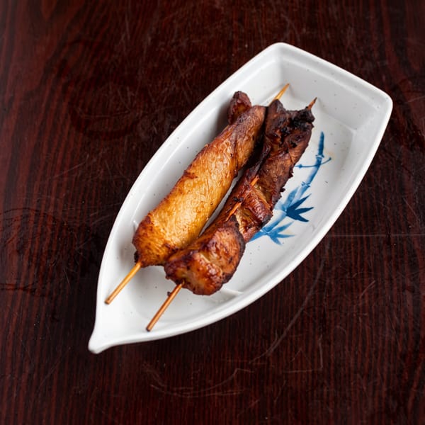 Two grilled skewers of chicken and tofu on a white boat-shaped plate with blue bamboo art.